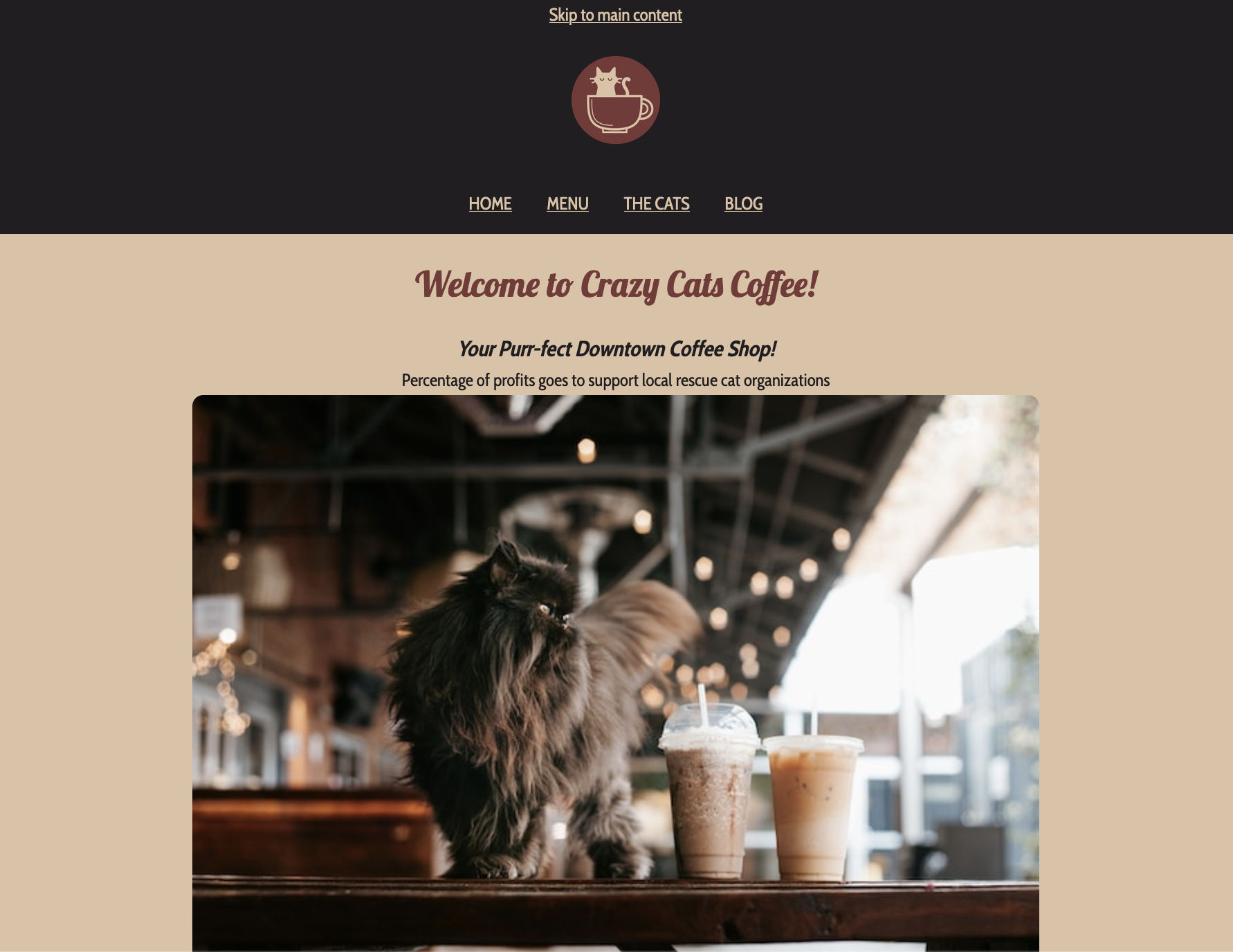 screenshot of Crazy Cats Coffee website. Colour theme is beige and brown. Top navigation is dark black-brown with beige text. It says Skip to main content. Underneath that there is the round red-brown logo of a beige cat sitting in a coffee cup. underneath there's links to HOME MENU and THE CATS. Below in red-brown calligraphy text is Welcome to Crazy Cats Coffee! The next line says in bold italic black text Your Purrfect Downtown Coffee Shop! underneath in regular weight and styled black text is Percentage of profits goes to support local rescue cat organizations. There's a picture of a dark brown long haired cat at coffee shop on a dark wood table by two iced coffees in plastic cups. Finally below the photo, in smaller red-brown calligraphy text than the welcome message is About Crazy Cats Coffee